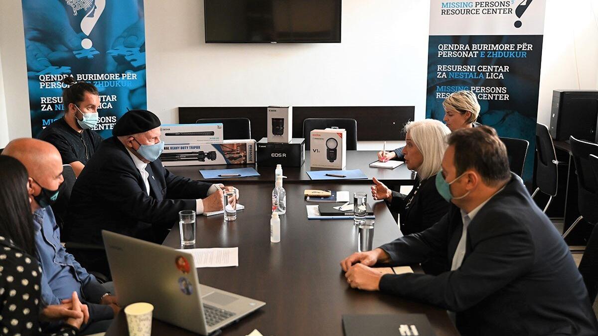 Several people seated around a conference table with laptops, notebooks, and water glasses. Behind them are banners reading “Missing Persons Resource Center” and boxes of equipment on the table.