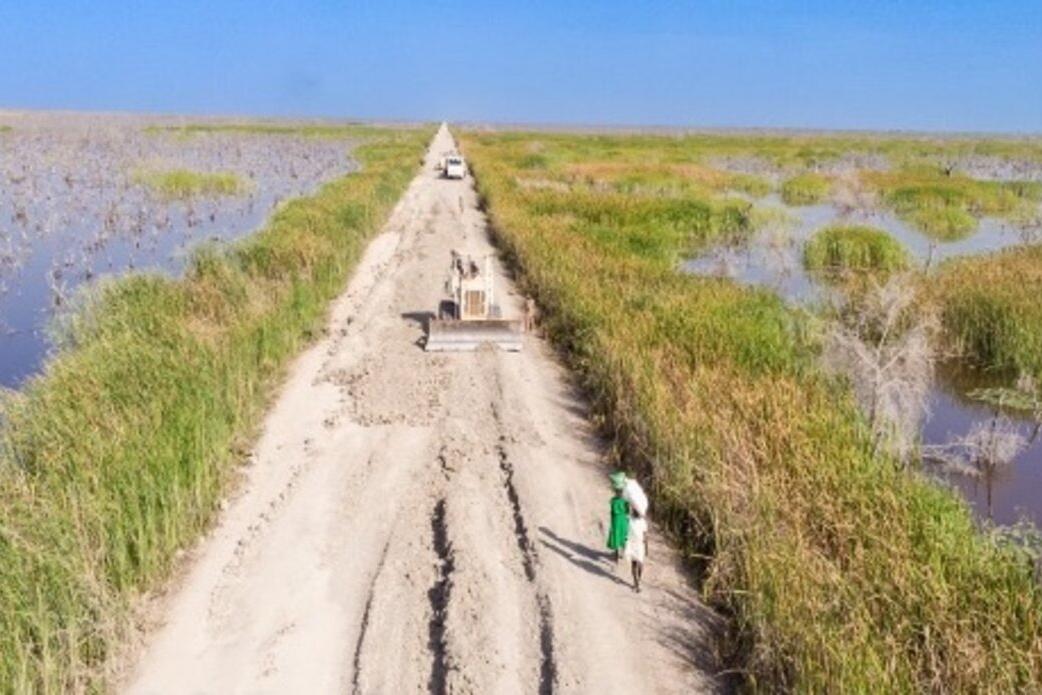 Maintaining the vital but often flood-affected road between Bentiu and Leer is a daunting task. Fortunately, our Pakistani engineers are up to it. An aerial view of a dirt road with grass and bodies of water on either side. On the road, there are some people walking.