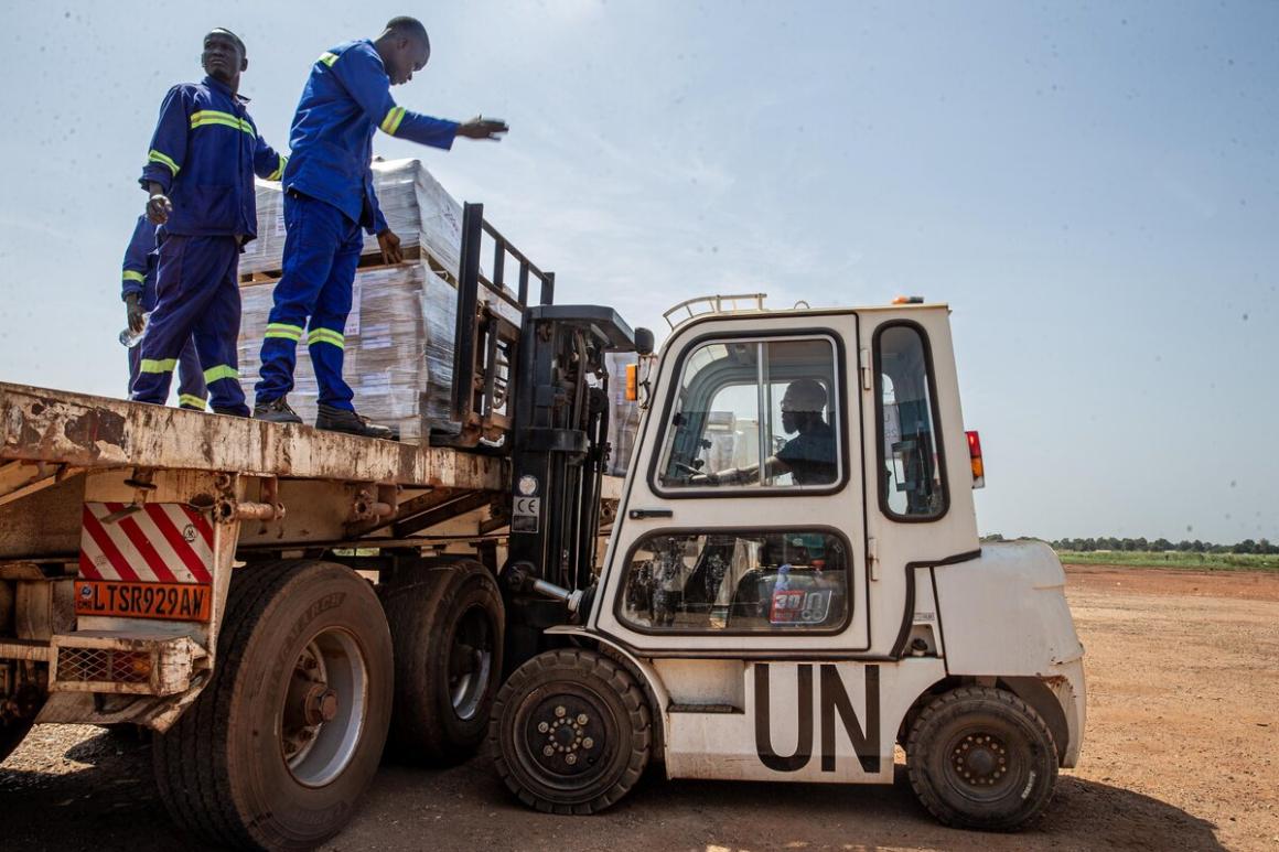 A forklift marked with “UN” is unloading large pallets wrapped in plastic from a flatbed truck. Two workers in blue uniforms with reflective stripes stand on the truck, guiding the process in an outdoor setting.